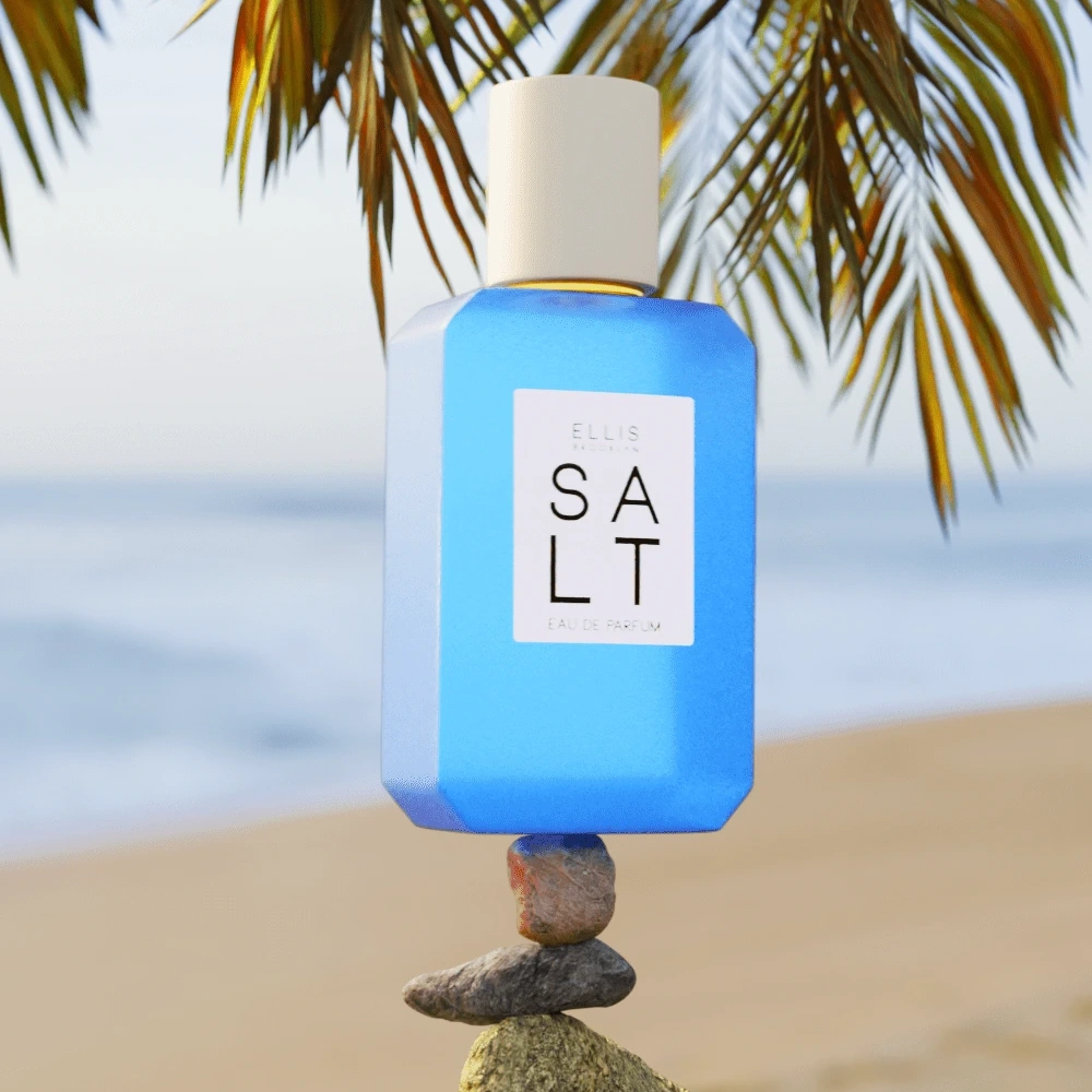 The perfume bottle is rectangular in shape, blue in color with a white label, on which in black font is written “Ellis Brooklyn SALT”. The flacon stands on a small stone balanced by another flat stone. The background depicts a sandy beach with eroded waves, partially covered by hanging green palm leaves.